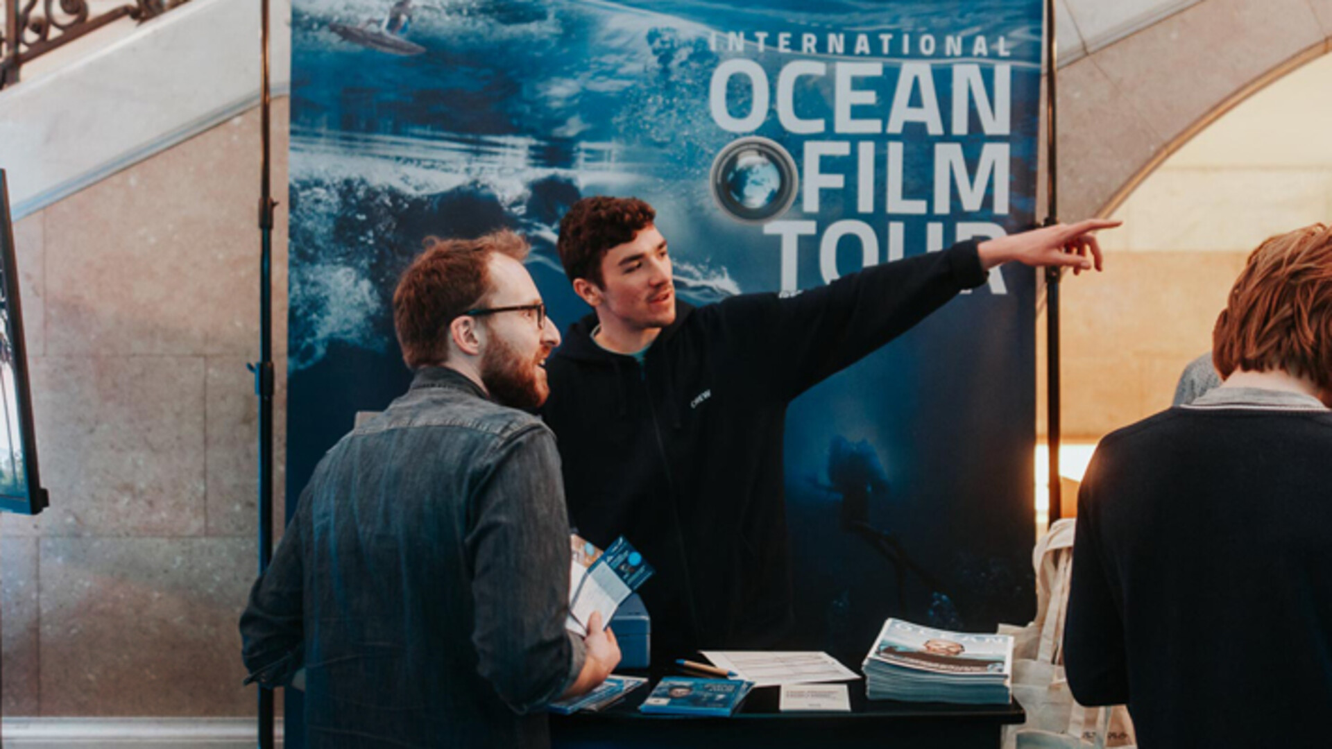 International Ocean Film Tour booth with staff providing information and handing out magazines to visitors.