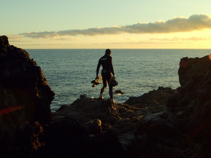 Diver in a wetsuit standing on rocks by the sea at sunset, holding diving gear in hand.