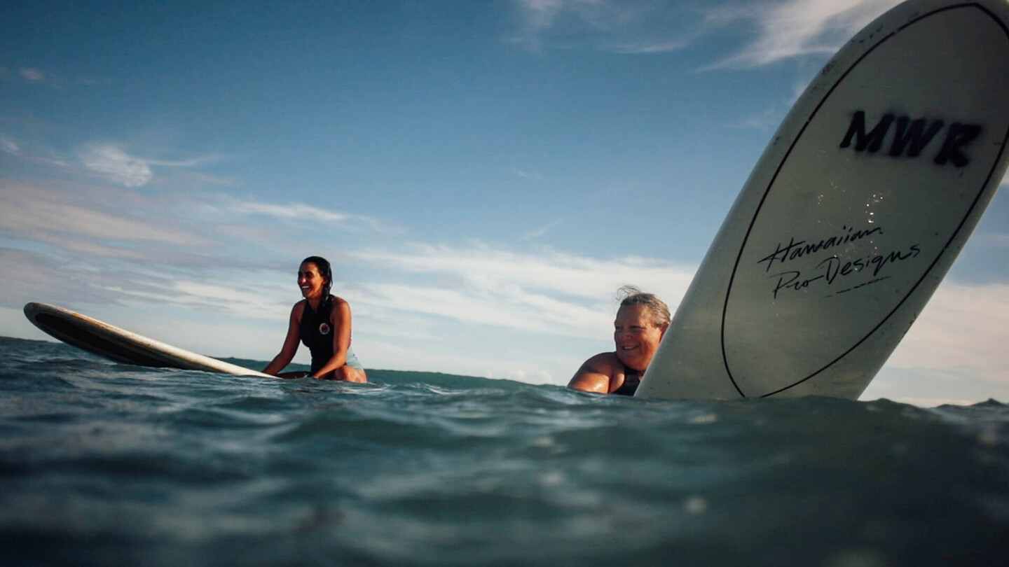 Two smiling female surfers sitting on their boards in the open sea, ready for the next wave.