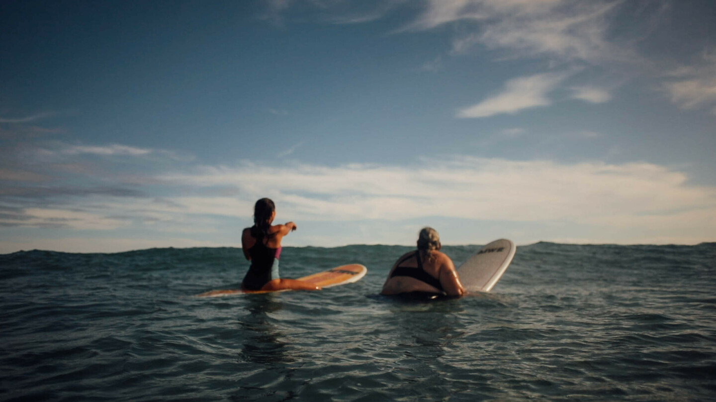 Two female surfers sitting on their boards in the open sea, looking at the approaching waves.